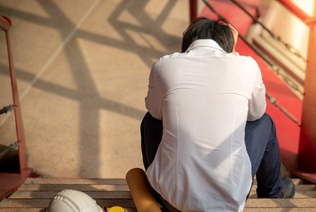 2 Un homme assis dans des escaliers à côté de son casque et de ses outils se tient la tête pour évoquer le Burnout professionnel