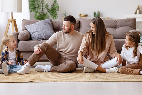 2 Photographie d'une famille assise sur le sol de leur salon. Le père, la mère et la grande sœur sont en train de regarder et sourire au petit garçon.