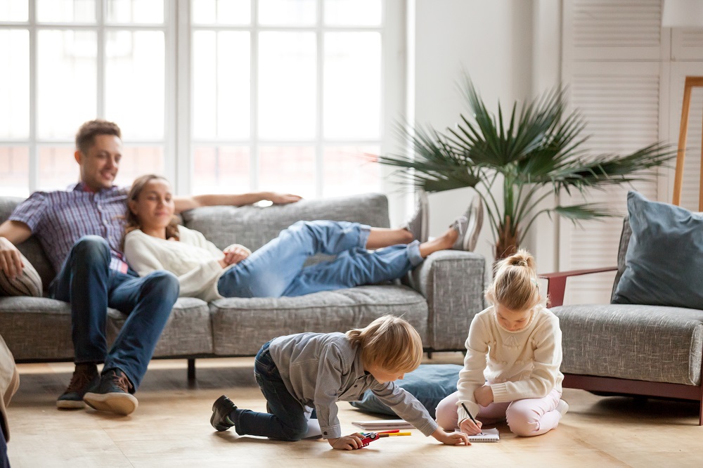 Une famille dans son salon avec deux parents qui se détendent sur le canapé et deux enfants qui jouent et dessinent