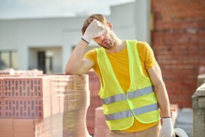 Un homme qui a chaud en gilet fluo enlève son casque de protection et touche son visage avec la main, se tenant près d'une pile de briques sur un chantier par une journée de fortes chaleurs.